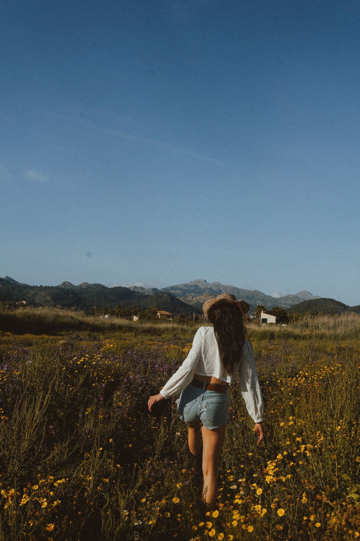 Woman in a straw hat walking through a wildflower meadow toward distant mountains